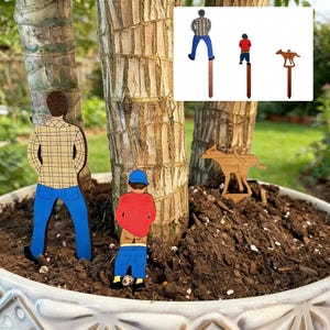 Puede incluir: Estacas de jardín de madera con figuras de personas y un perro. Las figuras están pintadas con detalles como camisas de cuadros, vaqueros azules y una camiseta roja. Las estacas se insertan en la tierra de una planta en maceta.
