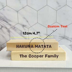 Puede incluir: Dos bloques de madera apilados uno encima del otro. El bloque superior tiene el texto "Hakuna Matata" y el bloque inferior tiene el texto "The Cooper Family". El bloque superior tiene 12 cm de ancho.