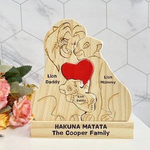 Puede incluir: Puzzle de madera de la familia del Rey Le&oacute;n con un coraz&oacute;n rojo en el centro. Las piezas del rompecabezas est&aacute;n etiquetadas como "Le&oacute;n Pap&aacute;", "Le&oacute;n Mam&aacute;" y "Le&oacute;n Beb&eacute;". La base del rompecabezas est&aacute; grabada con "Hakuna Matata La familia Cooper".