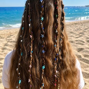 Peut inclure: Cheveux longs châtains coiffés de plusieurs tresses ornées de perles colorées. Les perles présentent des formes variées, notamment de petites étoiles de mer, des tortues marines et des breloques œil bleu, dans des tons bleu, rose, blanc et vert.