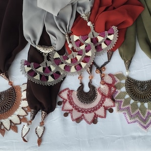 May include: An assortment of colorful scarves with decorative pendant necklaces. The scarves are in shades of brown, gray, red, and green. The necklaces feature intricate designs with beads and metal accents, showcasing a blend of textures and colors.