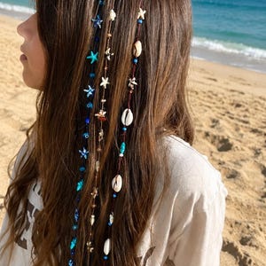 May include: Three hair wraps featuring a variety of decorative elements including small blue and white starfish charms, white cowrie shells, and assorted blue and brown beads strung along thin, dark-colored cords that hang down from the hair.