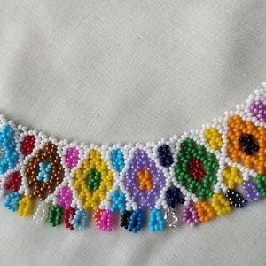 May include: A colorful beaded necklace with a floral design. The necklace features a white base with various colored beads, including pink, blue, yellow, and green. The necklace is made of small, round beads and has a gold-colored clasp.