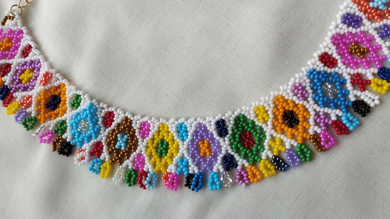 May include: A colorful beaded necklace with a floral design. The necklace features a white base with various colored beads, including pink, blue, yellow, and green. The necklace is made of small, round beads and has a gold-colored clasp.