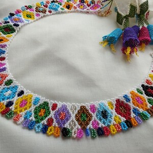 May include: A colorful beaded necklace with a white base and intricate patterns in various colors, including blue, yellow, green, and pink. The necklace has a gold clasp and is displayed on a white surface. The necklace is a piece of jewelry.