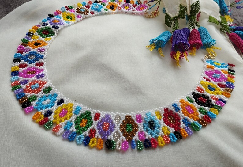 May include: A colorful beaded necklace with a white base and intricate patterns in various colors, including blue, yellow, green, and pink. The necklace has a gold clasp and is displayed on a white surface. The necklace is a piece of jewelry.