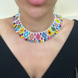 May include: A colorful beaded necklace with a white base and geometric patterns in shades of blue, yellow, orange, pink, and green. The necklace is worn on a person wearing a black top. The necklace is a statement piece.
