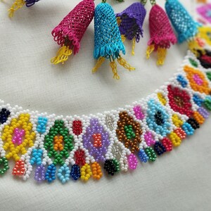 May include: A colorful beaded necklace with a white base, featuring intricate patterns in various colors like blue, yellow, and red. The necklace is adorned with dangling, bell-shaped accents in shades of pink, blue, and purple.