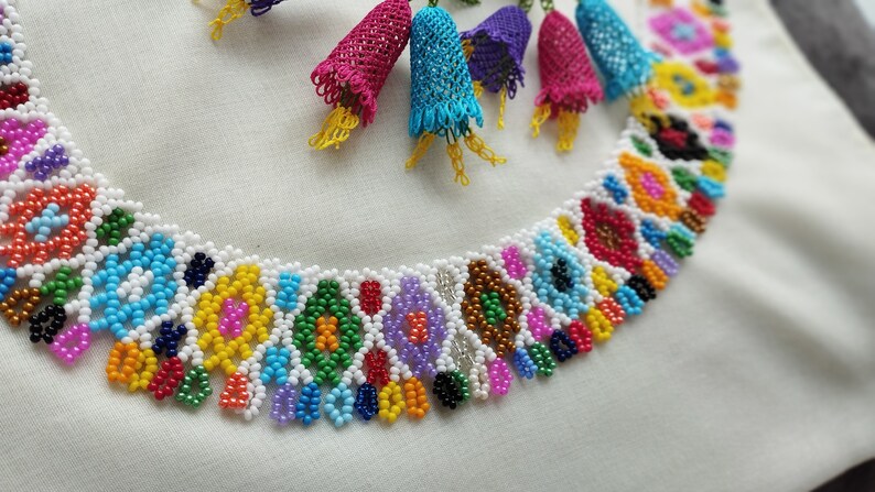 May include: A colorful beaded necklace with a white base, featuring intricate patterns in various colors like blue, yellow, and red. The necklace is adorned with dangling, bell-shaped accents in shades of pink, blue, and purple.