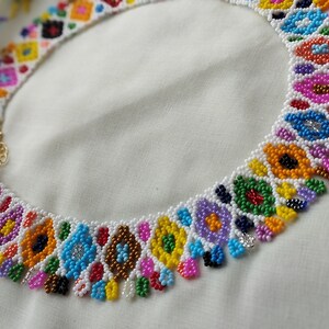 May include: A colorful beaded necklace with a floral design. The necklace features a variety of colors, including pink, blue, yellow, and green. The necklace is made of small beads and has a gold clasp. The necklace is on a white background.