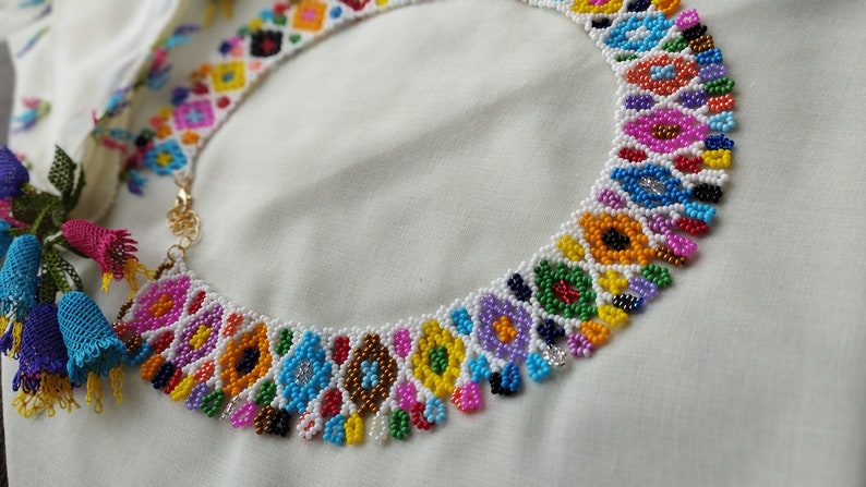 May include: A colorful beaded necklace with a floral design. The necklace features a variety of colors, including pink, blue, yellow, and green. The necklace is made of small beads and has a gold clasp. The necklace is on a white background.