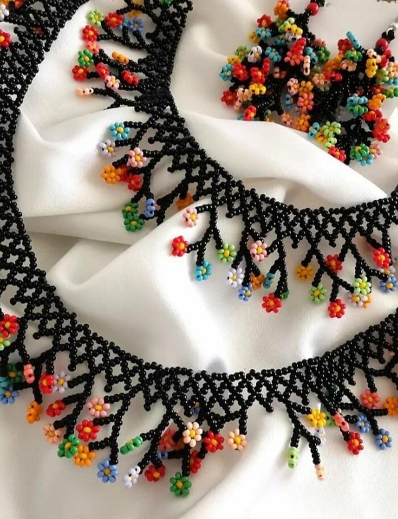 May include: A black beaded necklace with colorful flower accents. The necklace has a fringe design with small, colorful flowers hanging from the bottom. The necklace is paired with matching earrings.