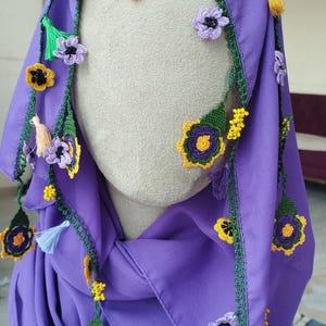 May include: A vibrant purple scarf adorned with crocheted floral accents. The flowers feature yellow, green, and purple details, with small tassels adding a decorative touch. The scarf is draped over a mannequin head.