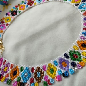 May include: A colorful beaded necklace with a floral design. The necklace features a variety of colors, including pink, blue, yellow, and green. The necklace is made of small beads and has a gold clasp.