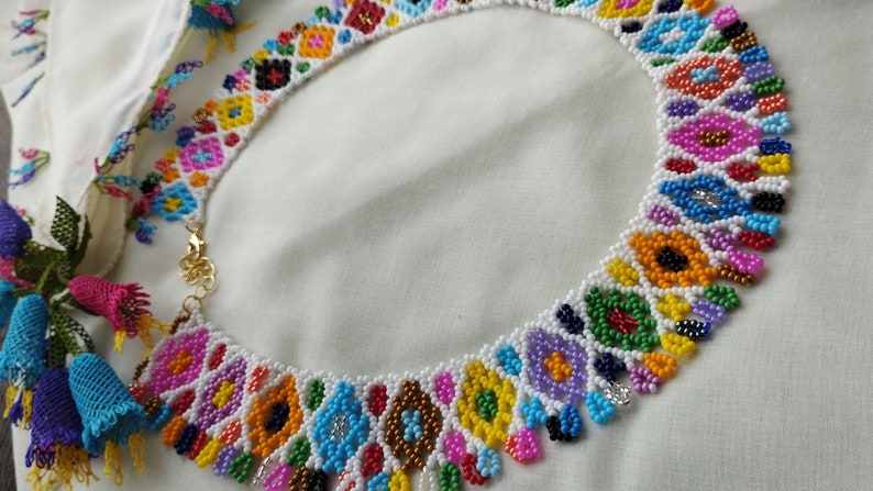 May include: A colorful beaded necklace with a floral design. The necklace features a variety of colors, including pink, blue, yellow, and green. The necklace is made of small beads and has a gold clasp.