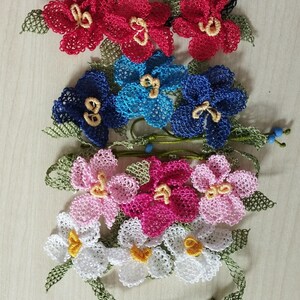 May include: Handmade floral bracelets in red, blue, pink, and white. Each bracelet features delicate, crocheted flowers with yellow centers and green leaves. The bracelets have black or green cords for tying.