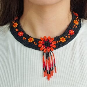 May include: A black beaded necklace with a large orange and black flower pendant. The necklace is made of small black beads and has a fringe of orange and black beads hanging from the pendant.