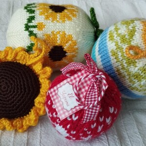 Könnte beinhalten: Vier gestrickte Ornamente, eines mit einem Sonnenblumenmuster, eines mit einem gestreiften Muster, eines mit einem roten Herzmuster und eines mit einer braunen Mitte und gelben Blütenblättern.