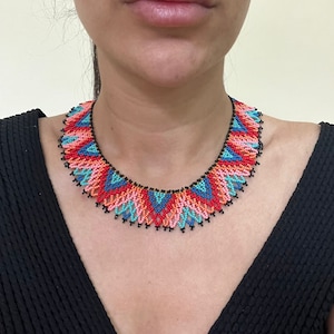 May include: A colorful beaded necklace with a geometric pattern in shades of red, orange, pink, blue, and black. The necklace is made of small beads and has a scalloped edge.