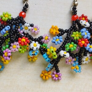 May include: A pair of dangle earrings with black beaded branches and colorful flower-shaped beads. The flowers are made of small, colorful beads in various shades of pink, blue, green, yellow, red, and white.