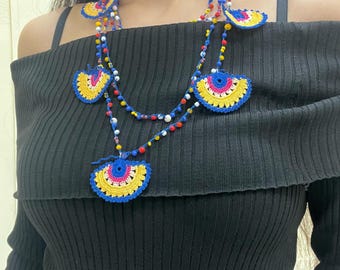 May include: A colorful crochet necklace featuring fan-shaped pendants in blue, yellow, and pink. The necklace has a beaded chain with red, white, and blue beads. The necklace is worn over a black off-the-shoulder top.