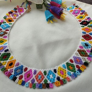 May include: A colorful beaded necklace with a geometric pattern. The necklace features a variety of colors, including white, pink, blue, yellow, and green. The necklace is made of small beads and has a detailed design.