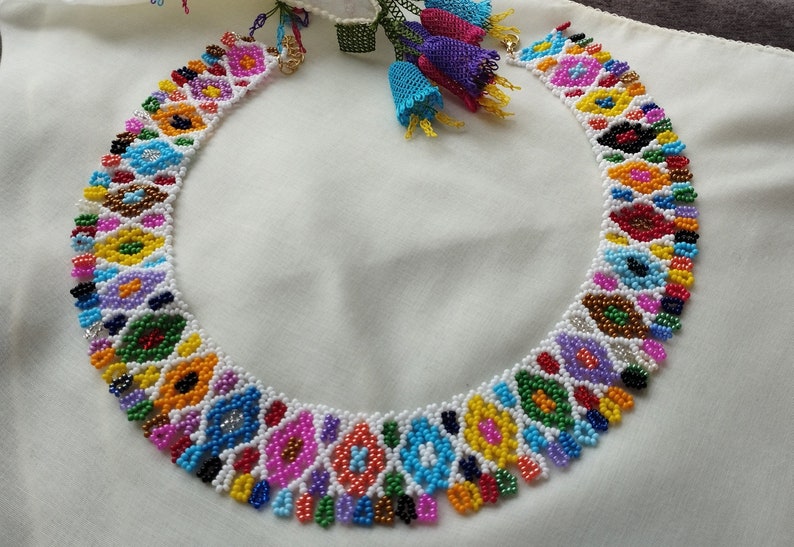 May include: A colorful beaded necklace with a geometric pattern. The necklace features a variety of colors, including white, pink, blue, yellow, and green. The necklace is made of small beads and has a detailed design.