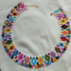 May include: A colorful beaded necklace with a floral design. The necklace features a variety of colors, including blue, yellow, pink, and green, set against a white background. The necklace has a gold-colored clasp and chain.