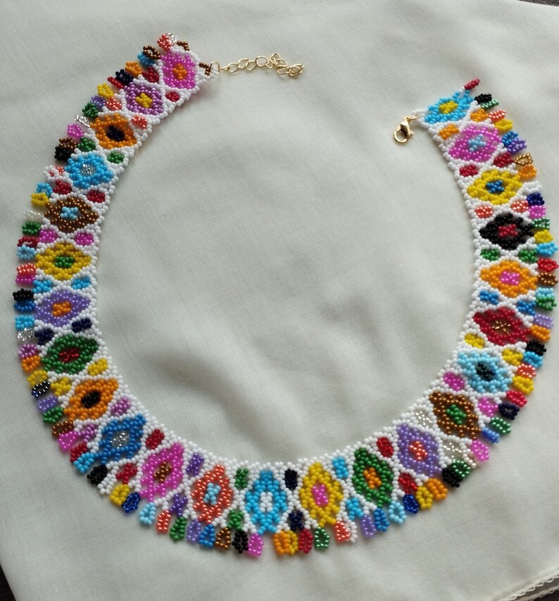 May include: A colorful beaded necklace with a floral design. The necklace features a variety of colors, including blue, yellow, pink, and green, set against a white background. The necklace has a gold-colored clasp and chain.