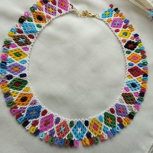 May include: A colorful beaded necklace featuring a repeating floral pattern in various colors including red, yellow, blue, and purple. The necklace is made with small beads and has a gold-colored clasp.