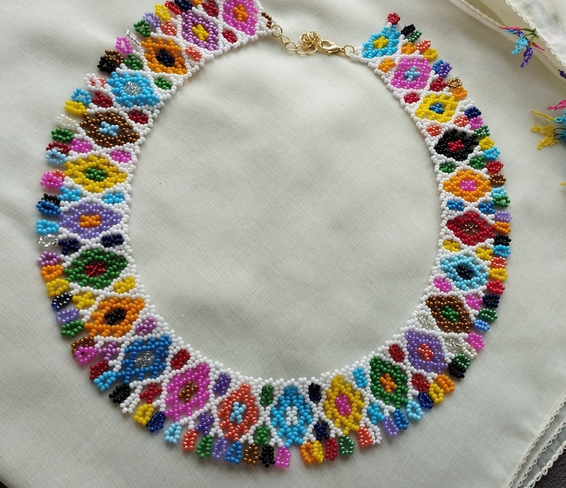 May include: A colorful beaded necklace featuring a repeating floral pattern in various colors including red, yellow, blue, and purple. The necklace is made with small beads and has a gold-colored clasp.