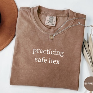 Puede incluir: Camiseta color espresso con el texto blanco "practicing safe hex". Un collar plateado con un pequeño colgante descansa sobre la camiseta. La camiseta está doblada y un sombrero marrón es visible en el fondo.