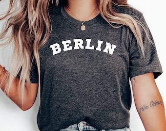 Pode incluir: Uma camiseta cinza escura com a palavra "BERLIN" impressa em branco, em arco sobre o peito. A camiseta é usada por uma pessoa com cabelo longo loiro e jeans azul.