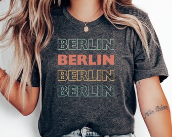 Pode incluir: Uma camiseta cinza escura com a palavra "BERLIN" repetida quatro vezes em um design empilhado. Cada palavra é contornada em uma cor diferente: verde, vermelho, amarelo e azul.