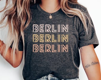 Pode incluir: Uma camiseta cinza escura com a palavra "BERLIN" repetida três vezes em um design empilhado. O texto está em uma fonte retrô com um esquema de cores rosa, amarelo e branco.
