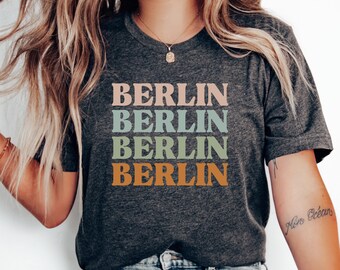 Pode incluir: Uma camiseta cinza escura com a palavra "BERLIN" repetida quatro vezes em um design empilhado. Cada palavra é de uma cor diferente: rosa, azul, verde e laranja.