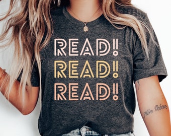 Puede incluir: Camiseta gris oscuro con la palabra "READ!" repetida tres veces en un diseño apilado. El texto está en una fuente retro con un degradado de colores de rosa claro a amarillo a melocotón. La camiseta es de cuello redondo.