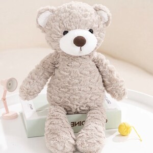 Könnte beinhalten: Ein plüschiger grauer Teddybär mit weißem Gesicht und brauner Nase. Der Bär sitzt auf einem grünen Buch mit einem gelben Wollknäuel daneben.