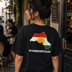 Può includere: Una t-shirt nera a maniche corte con una grafica sul retro. La grafica mostra il contorno di una mappa riempito con le strisce orizzontali rossa, bianca e verde della bandiera del Kurdistan, un emblema solare giallo e il testo KURDISTAN.