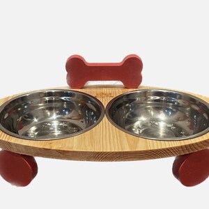 Könnte beinhalten: Hunde-Futternapf-Ständer aus hellem Holz mit zwei Edelstahlnäpfen. Der Ständer hat rote Akzente, darunter ein knochenförmiges Detail und abgerundete Füße. Die Schalen sind oval und für Tierfutter oder Wasser bestimmt.