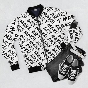 May include: A white bomber jacket with a black zipper and black trim. The jacket is covered in a repeating pattern of the words "Make Art" in black. The jacket is laid out on a white surface with a pair of black jeans and black and white sneakers.