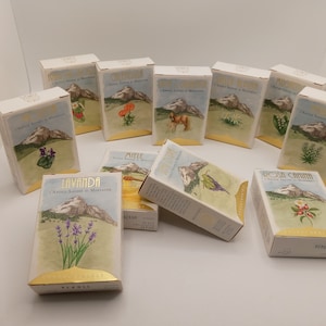 Può includere: Una collezione di scatole rettangolari con illustrazioni di montagne e piante. Ogni scatola presenta una pianta diversa, come lavanda, calendula e rosa. Le scatole hanno dettagli dorati e testo in italiano.