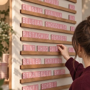May include: A wall-mounted wooden menu board with horizontal rails holding pink letter tiles. The text reads ICE CREAM & GELATO MENU, CHERRY RIPPLE, CLASSIC VANILLA, BELGIAN CHOCOLATE, PISTACHIO PERFECT, MINT CHOC CHIP, SALTED CARAMEL, STRAWBERRY SWIRL, HONEY SORBET, FROZEN YOGURT.
