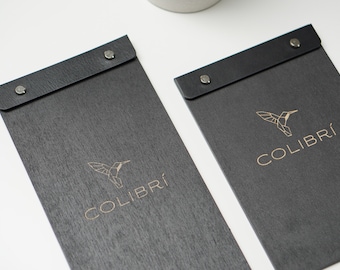 Puede incluir: Dos cubiertas de menú de cuero negro con un logotipo de colibrí dorado y la palabra "COLIBRÍ" impresa en cada una.