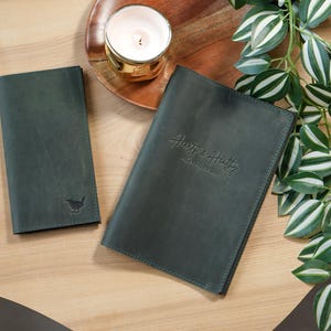 May include: A dark green leather notebook cover and a matching slim wallet. The notebook cover features the embossed text "Heart & Healthy" and the wallet displays a small bird emblem. Both items have visible stitching along the edges.