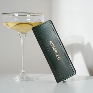 May include: A green leather table card with gold lettering that reads "RESERVED". The card is sitting on a white surface next to a champagne flute.