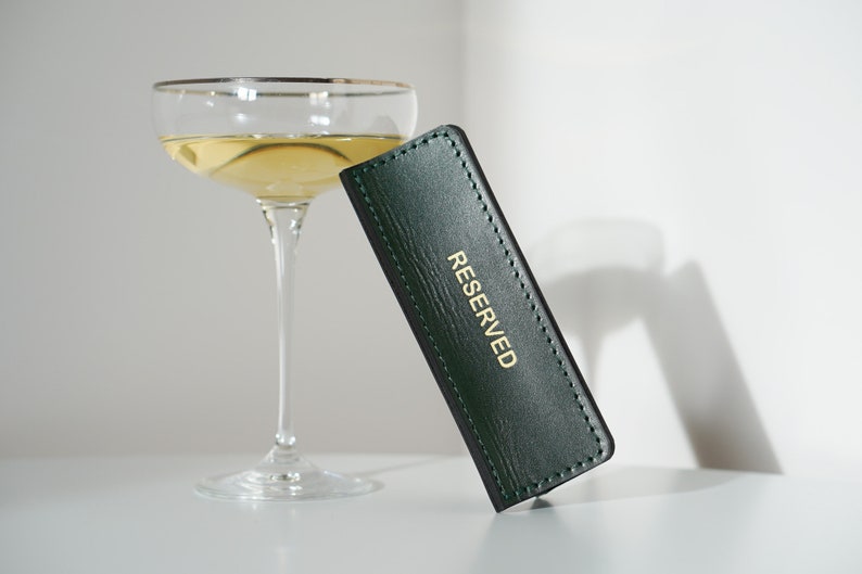 May include: A green leather table card with gold lettering that reads "RESERVED". The card is sitting on a white surface next to a champagne flute.