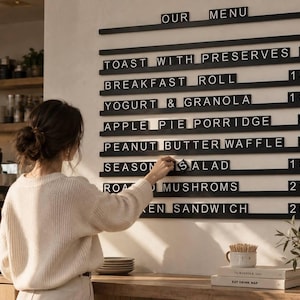 May include: A wall-mounted black menu board with horizontal rails holding white letter tiles. The text reads: OUR MENU, TOAST WITH PRESERVES 8, BREAKFAST ROLL 15, YOGURT & GRANOLA 16, APPLE PIE PORRIDGE 5, PEANUT BUTTER WAFFLE 3, SEASONAL SALAD 18, ROASTED MUSHROOMS 21, CHICKEN SANDWICH 25.