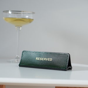 May include: A green leather table sign with gold stitching and the word "RESERVED" in gold lettering.
