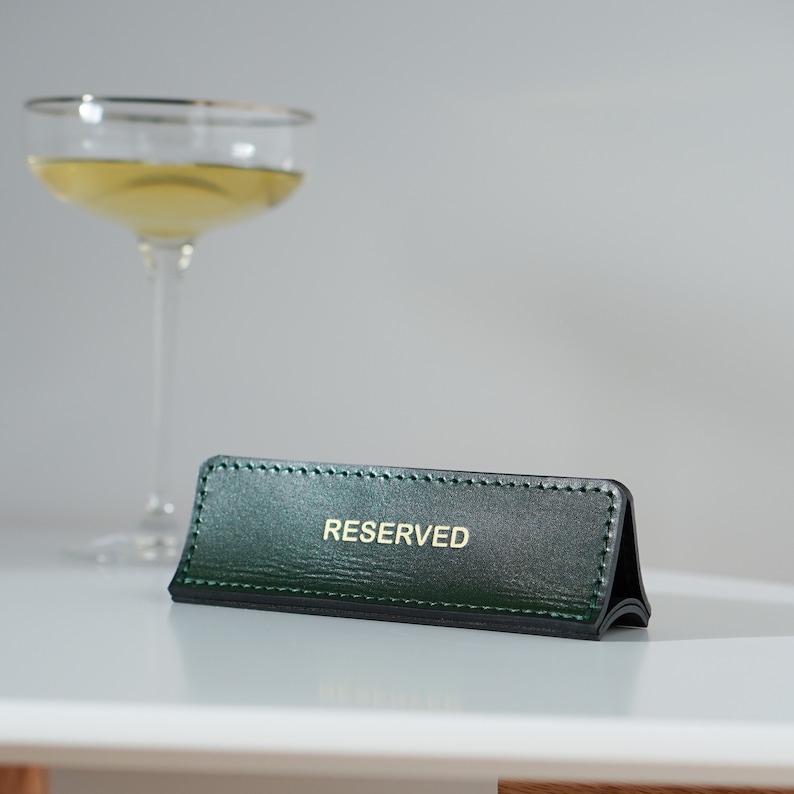 May include: A green leather table sign with gold stitching and the word "RESERVED" in gold lettering.